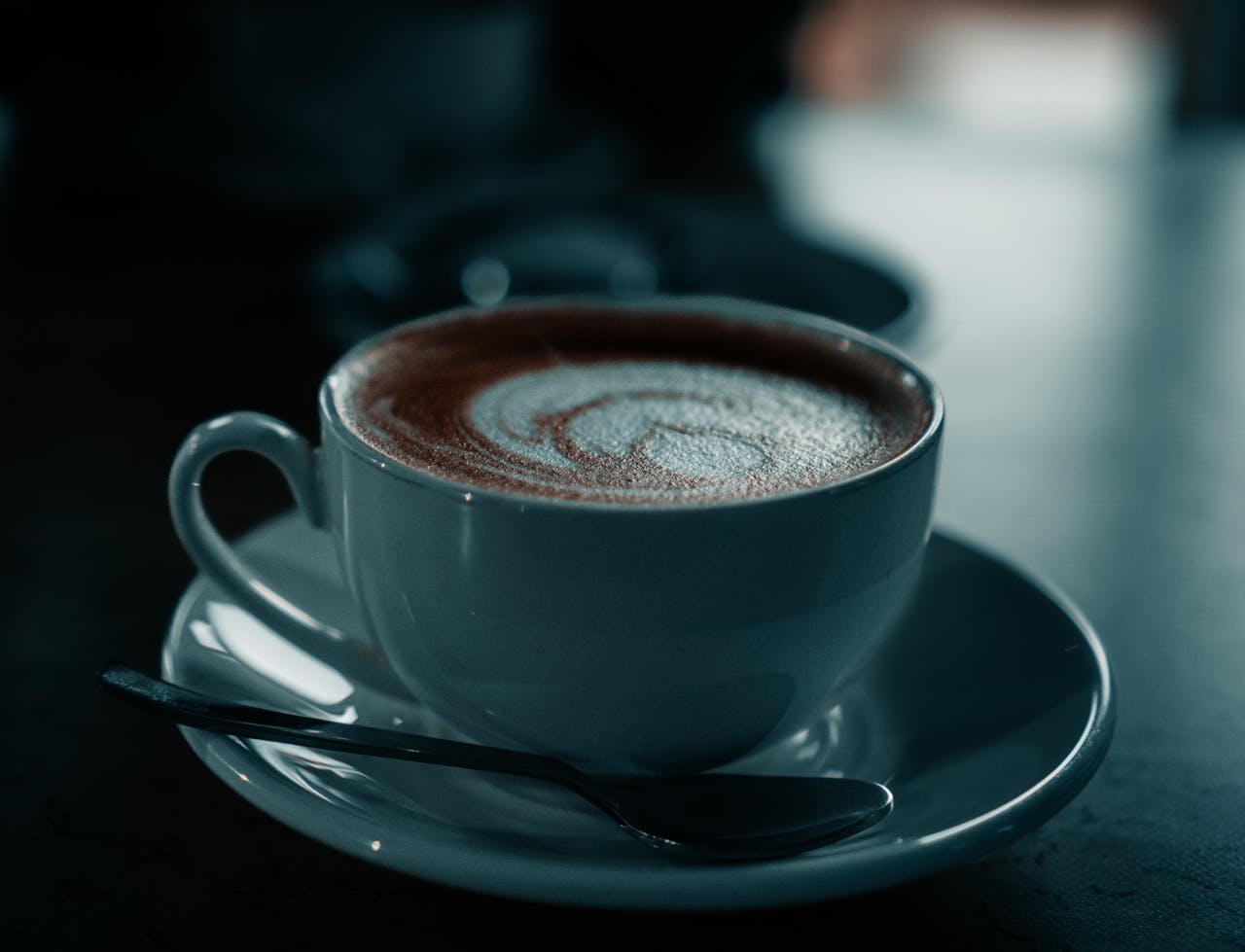A serene close-up of a cappuccino cup with a spoon on a saucer in a moody setting.